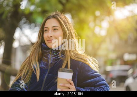 Ragazza latina che beve un caffè seduto su una panchina in un parco pubblico con spazio fotocopie. Foto Stock