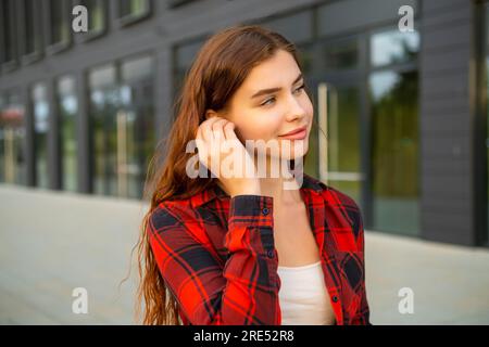 Vicino al business center, una giovane donna che indossa una camicia rossa a scacchi mette le cuffie nelle orecchie Foto Stock