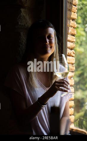 Bella giovane donna con lunghi capelli biondi sorridenti e guardando la macchina fotografica che tiene in mano un bicchiere di vino bianco illuminato da una luce soffusa proveniente da un vento Foto Stock