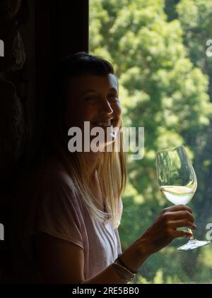 Giovane, graziosa, donna alternativa con i capelli lunghi e un naso che traspira retroilluminato con un bicchiere di vino bianco in mano illuminato da una luce soffusa Foto Stock