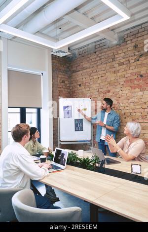 Un manager di sesso maschile che parla con i colleghi nella sala riunioni mentre si trova vicino a una lavagna a fogli mobili con delle tabelle. La startup, il coworking, il manager Foto Stock