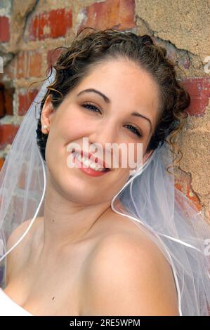 La sposa trasmittente si appoggia contro un muro di mattoni rustici. Lei è sorridente e felice. Indossa un abito e un velo senza spalline. Foto Stock