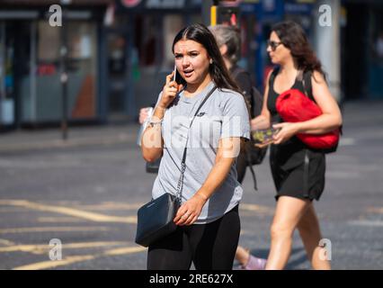 Giovane donna che cammina in una zona urbana trafficata e parla su un cellulare o cellulare, in estate in Inghilterra, Regno Unito. Foto Stock