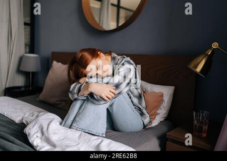 Vista frontale della donna rossa millenaria depressa seduta sul letto abbraccio ginocchia perse in cattive riflessioni pessimistiche soffrono da sola. Giovane donna stressata Foto Stock