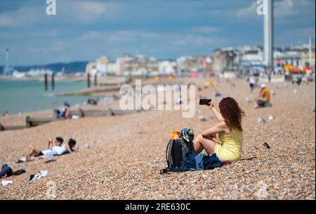 Donna che scatta selfie con lo smartphone, seduto sulla spiaggia di Brighton in una nuvolosa giornata estiva a Brighton & Hove, East Sussex, Inghilterra, Regno Unito. Foto Stock