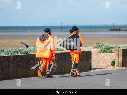 Un paio di lavoratori maschi, vestiti ad alta visibilità, passeggiate lungo la strada lungomare, raccolta di rifiuti dai bidoni della spazzatura e percorso di pulizia sul lungomare in estate, Regno Unito. Foto Stock