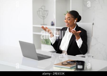 Allegra donna d'affari afroamericana in abbigliamento formale che effettua videochiamate su un computer portatile, lavora in interni leggeri di un ufficio Foto Stock