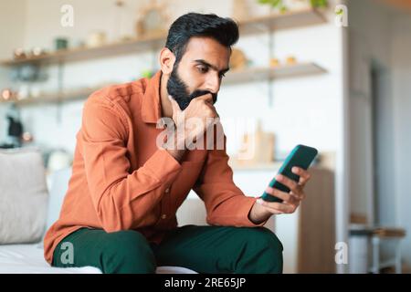 Un indiano premuroso che tiene in mano messaggi e pensieri da smartphone a casa Foto Stock
