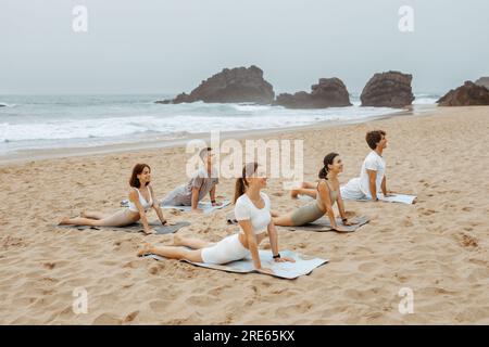 Tranquillità delle maree. Gruppo di giovani uomini e donne che praticano yoga e fanno posa di cobra sui tappetini, esercitandosi sulla spiaggia Foto Stock