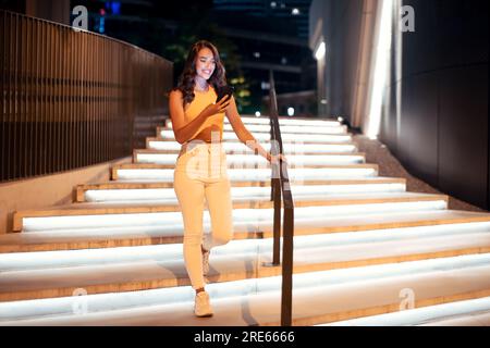Foto completa di una donna che usa il telefono durante la passeggiata notturna in città, scendendo le scale illuminate, spazio libero Foto Stock