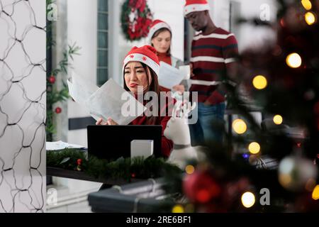 Dipendente sdepresso del lavoro turbato dallo stressante lavoro d'ufficio dell'azienda in ritardo a Natale. Stressato il lavoro dei dipendenti in un luogo di lavoro decorato per le feste, cercando di portare a termine un progetto difficile Foto Stock