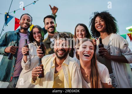Ritratto di un grande gruppo di amici felici e giovani che sorridono e si divertono a festeggiare una festa sul tetto, una riunione sociale o un incontro di compleanno di sera, bevendo birra e divertendosi insieme. Foto di alta qualità Foto Stock