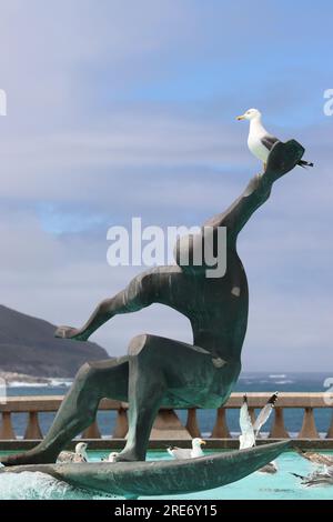 Un gabbiano arroccato sulla cima di una statua di surfista dello scultore spagnolo José Castiñeiras Iglesias, nella Fontana di Los Surfistas, sulla spiaggia di Orzán, la Coruna, Spagna. Foto Stock