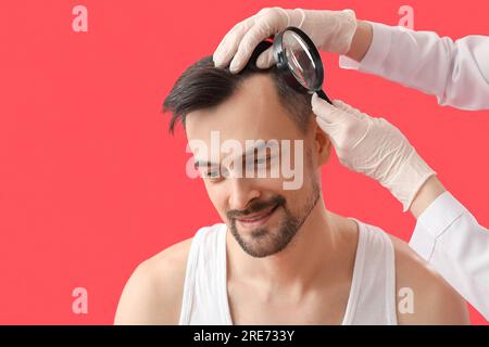 Medico che esamina i capelli del giovane con lente di ingrandimento su sfondo rosso, primo piano Foto Stock