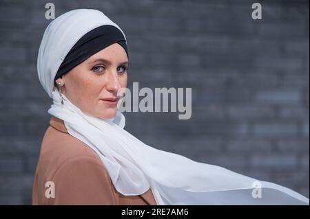 Ritratto di una giovane donna dagli occhi blu in un hijab contro un muro di mattoni grigi. Sciarpa bianca fluttua nel vento. Foto Stock