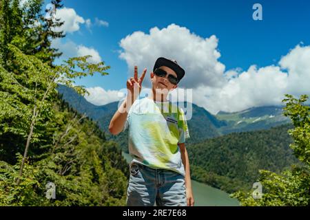 Ritratto del viaggiatore escursionistico ragazzo caucasico che solleva due dita sulla mano mentre la pace gesture.beautifulturquois montagna blu lago sullo sfondo Foto Stock