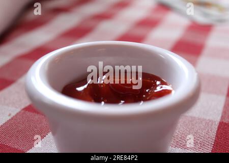 Una foto ravvicinata di una piccola pentola di ketchup di pomodoro su un tavolo a scacchi rosso e bianco. Foto Stock