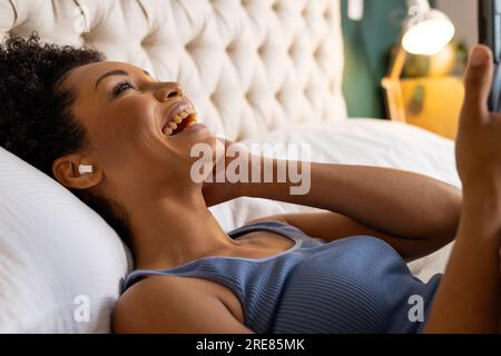 Donna birazziale felice con gli auricolari sdraiati sul letto e che usa lo smartphone in camera da letto Foto Stock