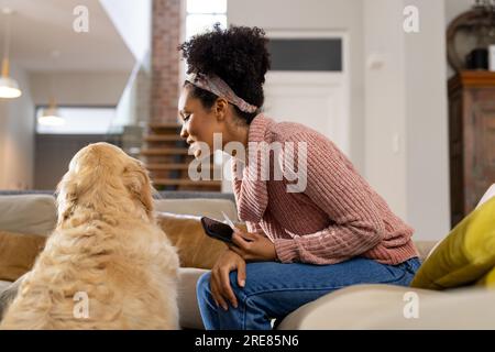 Felice donna birazziale che accarezza il cane e usa lo smartphone a casa Foto Stock