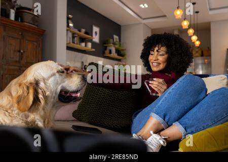 Felice donna birazziale che accarezza il cane e beve tè a casa Foto Stock