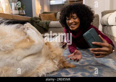 Felice donna birazziale che accarezza il cane e usa lo smartphone a casa Foto Stock
