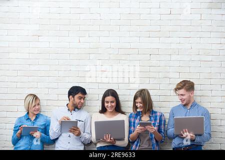 Amici allegri che guardano video divertenti sul notebook mentre sono distratti dalla navigazione in Internet con l'aiuto dei loro gadget, parete bianca su sfondo Foto Stock