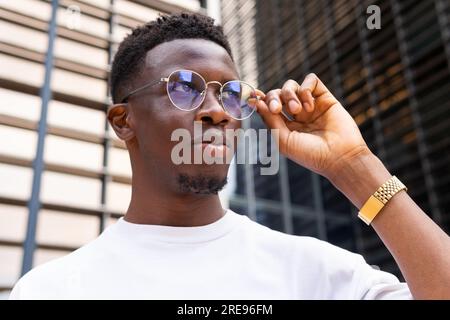 Serio uomo afro-americano con outfit alla moda e occhiali da vista che guardano lontano mentre si trovano contro edifici moderni Foto Stock
