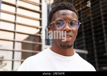 Serio uomo afro-americano con outfit alla moda e occhiali da vista che guardano la macchina fotografica mentre si trovano contro edifici moderni Foto Stock
