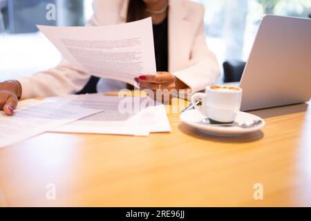 Anonima donna d'affari afroamericana con occhiali e abiti formali seduta al tavolo vicino al computer portatile e una tazza di caffè mentre leggeva i documenti in ufficio Foto Stock