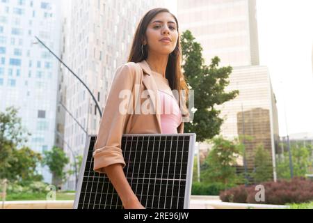 Dal basso di una donna imprenditrice in blazer in piedi per strada con pannello solare portatile e guardando la fotocamera Foto Stock