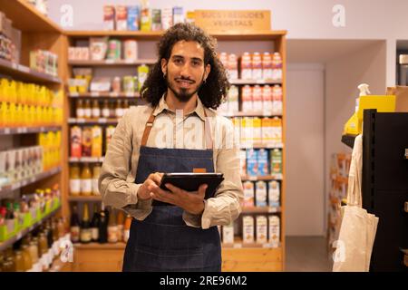 Ritratto di un lavoratore etnico maschio felice con lunghi capelli ricci in abiti casual in piedi vicino agli scaffali che guarda la fotocamera sorridente mentre lavora su un tablet Foto Stock