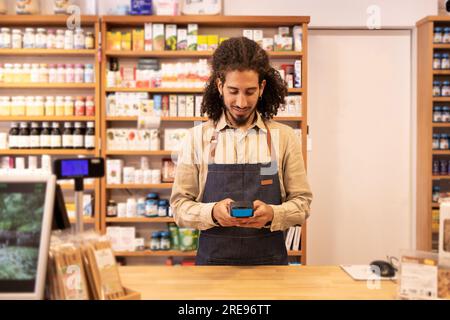 Sorridente maschio etnico felice con i capelli ricci in abiti casual in piedi su un tavolo di legno mentre si gestiva ed elaborava il pagamento finanziario sulla macchina POS i Foto Stock