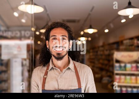 Ritratto di un uomo barbuto positivo in grembiule sorridente e che guarda la macchina fotografica mentre si trova vicino al buio sfondo illuminato sfocato nel supermercato Foto Stock