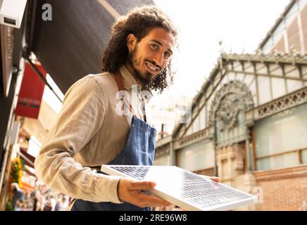 Angolo basso di giovani uomini etnici positivi in abiti casual in piedi per strada e guardando la fotocamera mentre si tiene il pannello solare portatile alla luce del giorno Foto Stock