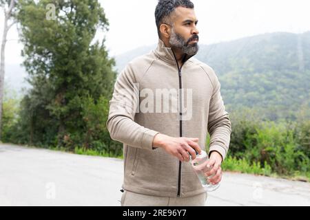 Uomo maturo barbuto in abbigliamento sportivo con una bottiglia d'acqua in una strada rurale Foto Stock