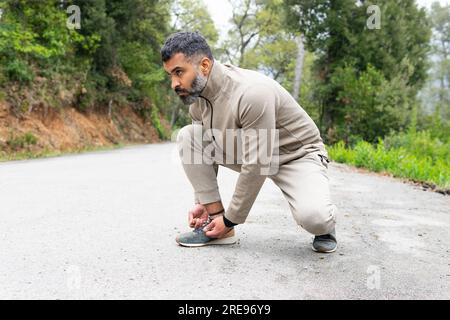 Vista laterale di un atleta indiano barbuto con abbigliamento sportivo che guarda lontano mentre si siede su asfalto e si lega un paio di scarpe da ginnastica vicino a alberi verdi Foto Stock