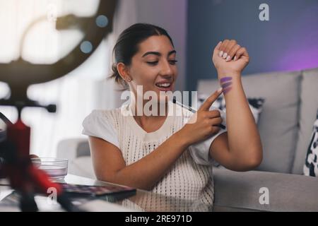Sorridente giovane influencer ispanica che punta al polso con toni viola di ombre oculari mentre parla con la videocamera per registrare video a casa Foto Stock
