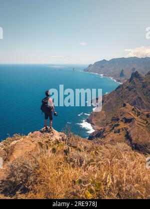 Vista posteriore di un anonimo escursionista maschile con zaino e macchina fotografica in piedi sulla scogliera mentre ammiri il paesaggio marino di Tenerife in Spagna alla luce del giorno Foto Stock