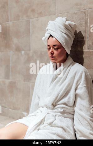 Una giovane donna in accappatoio bianco sulla testa mentre si siede con gli occhi chiusi in un hammam piastrellato durante le vacanze Foto Stock