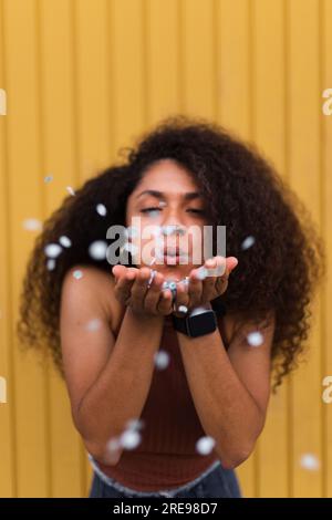Giocosa donna afroamericana con capelli afro soffia confetti mentre festeggiamo le vacanze in città e divertitevi Foto Stock