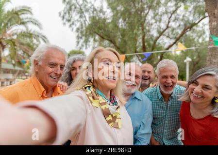 Donna sorridente di mezza età con i capelli biondi che si autoritratta con gli amici durante le celebrazioni all'aperto nel parco Foto Stock