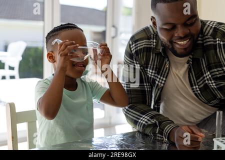 Felice padre e figlio afro-americani che fanno esperimenti di chimica insieme nel soleggiato soggiorno Foto Stock