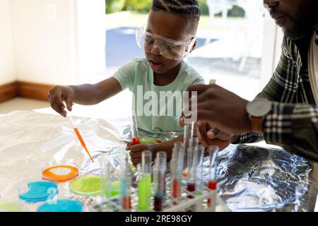 Felice padre e figlio afro-americani che fanno esperimenti di chimica insieme nel soleggiato soggiorno Foto Stock