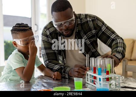 Felice padre e figlio afro-americani che fanno esperimenti di chimica insieme nel soleggiato soggiorno Foto Stock