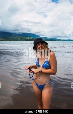 Giovane e allegra turista che indossa bikini blu e cappello panama in piedi sulla spiaggia vicino a salutare il mare contro le montagne tenendo in mano una macchina fotografica d'epoca durante la Sum Foto Stock