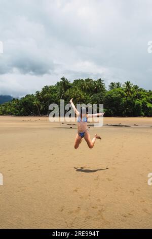 Tutto il corpo di una giovane e felice turista in bikini e cappello che salta con le braccia alzate sulla spiaggia sabbiosa di Uvita contro gli alberi tropicali in Costa Rica Foto Stock