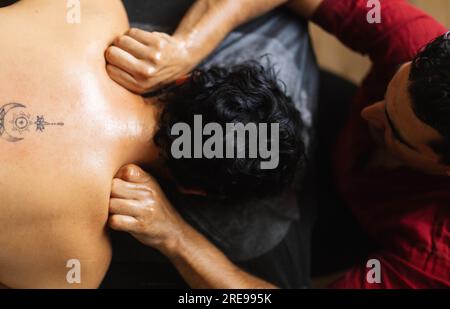 Vista dall'alto del terapista maschile mentre si è seduti alla scrivania e si impastano le spalle con le dita piegate su una cliente femminile anonima Foto Stock