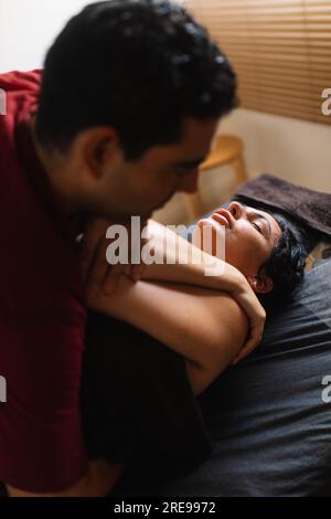 Vista laterale del massaggiatore ispanico adulto che guarda in basso mentre esercita pressione sul braccio incrociato di una giovane donna etnica sdraiata con gli occhi chiusi per alleviare lo stress Foto Stock