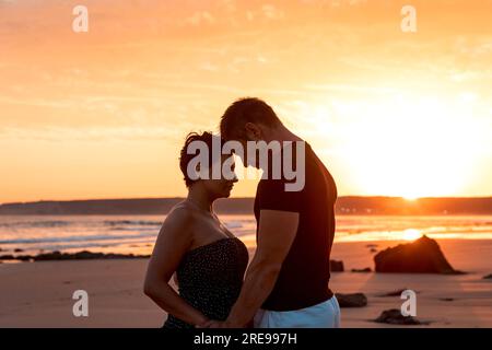 Vista laterale dell'amorevole marito e moglie che si tengono per mano mentre si stendono sulla costa sabbiosa vicino al mare che sventola contro il cielo al tramonto Foto Stock