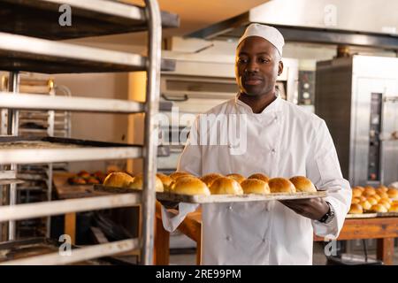 Un fornaio afro-americano felice nella cucina del forno con vassoio da forno e panini freschi Foto Stock
